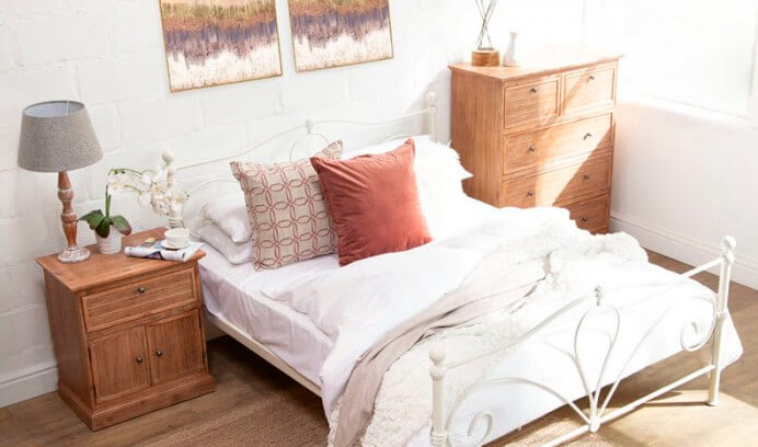 White metal bed with white bedding and one acacia wood pedestal one side of the bed and on the other side an acacia wood chest of drawers